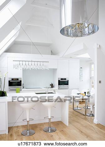 Stock Image - White Bomba stools at breakfast bar in modern white loft conversion kitchen with wooden flooring. Fotosearch