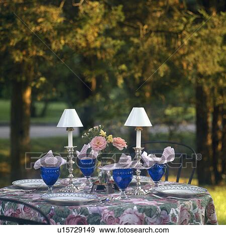 Stock Photo - White candle-lamps on table set for dinner with blue glasses and rose-patterned cloth in country garden. Fotosearch