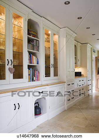 Stock Photo - White fitted cupboards with lighting and glass doors in townhouse kitchen. Fotosearch