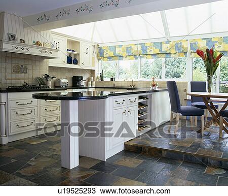 Stock Image - White fitted unit dividing kitchen from split-level dining area in modern extension with slate-tiled floor. Fotosearch