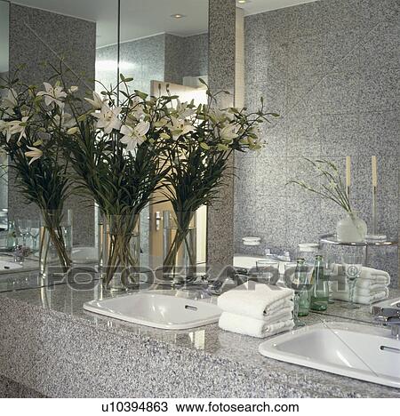 Stock Image - White lilies on grey granite vanity units with double white basins below mirror wall in modern bathroom. Fotosearch
