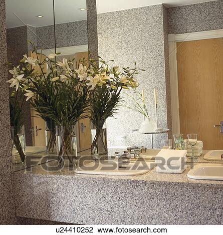 Stock Image - White lilies on grey granite vanity units with double white basins below mirror wall in modern bathroom. Fotosearch