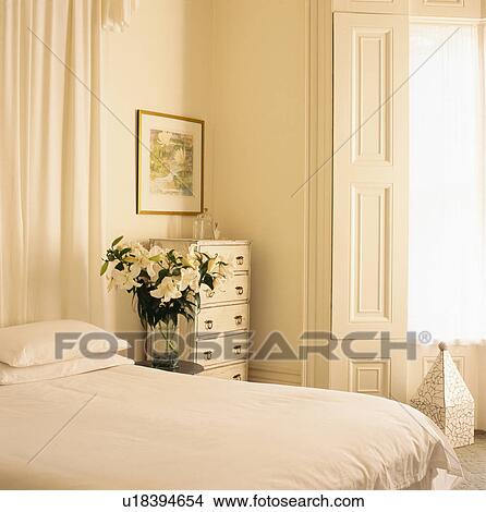 Picture - White lilies on table beside bed with white linen in light and airy bedroom with original white shutters and the window. Fotosearch