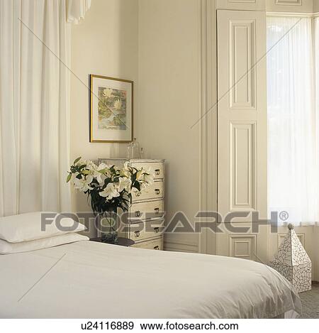 Stock Photo - White lilies on table beside bed with white linen in light and airy bedroom with original white shutters and the window. Fotosearch
