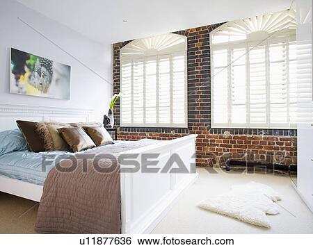 Stock Photograph - White plantation shutters on window in exposed brick wall in modern bedroom with pastel blue and brown bedlinen. Fotosearch