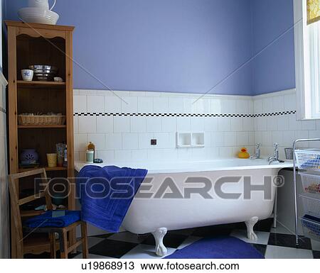 Stock Image - White tiled wall above rolltop bath in blue bathroom with freestanding wooden shelves. Fotosearch