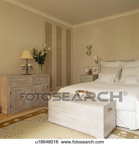 Stock Photograph - White wooden chest below bed with white linen in modern white bedroom. Fotosearch