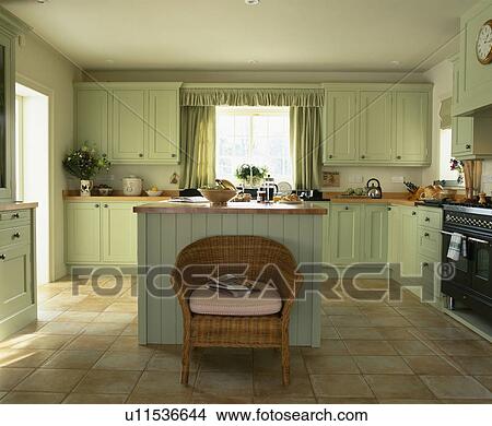 Picture - Wicker chair in front of island unit in kitchen with limestone floor and pastel gren fitted cupboards. Fotosearch