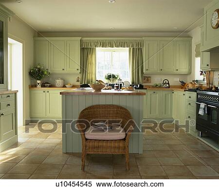 Stock Photography - Wicker chair in front of island unit in kitchen with limestone floor and pastel green units. Fotosearch
