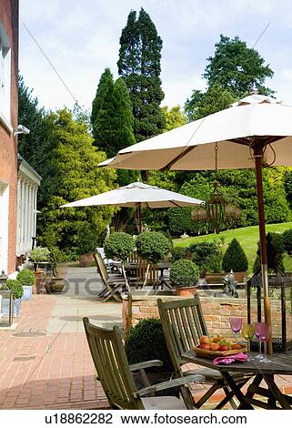 Stock Image - Wooden chairs and tables below white parasols on brick patio in large country garden. Fotosearch