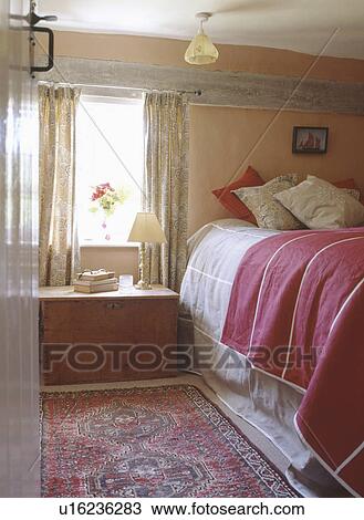 Stock Image - Wooden chest below window in cottage bedroom with cushions and red throw on the bed. Fotosearch