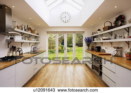Stock Image - Wooden flooring and glass patio doors in modern kitchen extension with fitted white units. Fotosearch