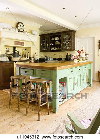 Stock Image - Wooden stools at green island unit in pale yellow country kitchen with wooden flooring. Fotosearch