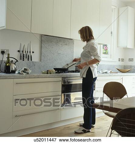Stock Photo - Young woman cooking at stove in modern white kitchen. Fotosearch