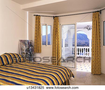 Stock Photography - Bed with striped bedlinen in modern Spanish bedroom with patio doors onto balcony&#10;. Fotosearch