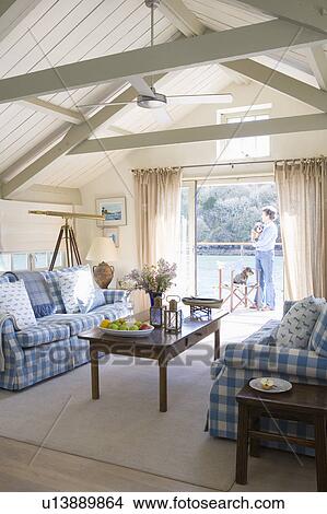 Picture - Blue+white checked sofas in living room in converted coastal boathouse with view of woman standing on balcony. Fotosearch