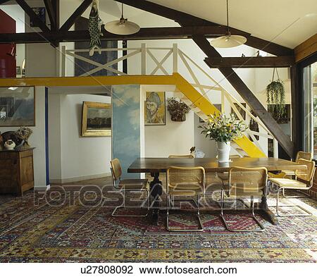 Stock Image - Chrome and cane chairs at long wooden table in loft conversion dining room. Fotosearch