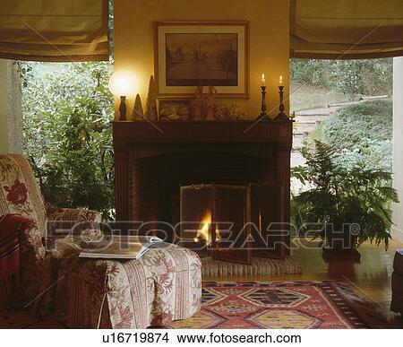 Picture - Comfy armchair and stool in front of fireplace with lighted fire between large windows in cosy living room. Fotosearch