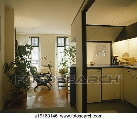 Stock Photograph - Compact fitted kitchen built into corner of large apartment living room with parquet flooring. Fotosearch