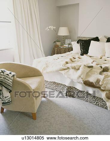 Stock Photography - Cream tub-chair in modern bedroom with faux fur throws on bed. Fotosearch
