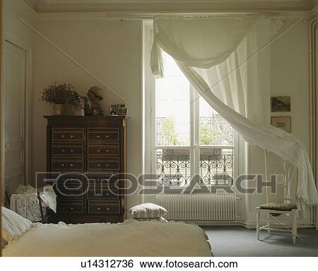 Stock Photograph - Cream voile curtains at French windows in white traditional townhouse bedroom. Fotosearch