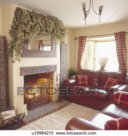 Dried hops on mirror above stone fireplace & metal fireguard in cream cottage living room with brown leather sofas View Large Photo Image Stock Photography - Dried hops on mirror above stone fireplace & metal fireguard in cream cottage living room with brown leather sofas. Fotosearch