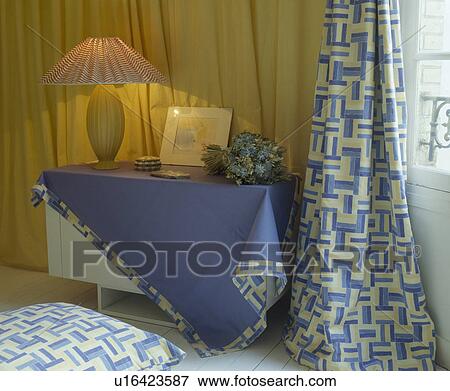 Stock Photo - Lighted lamp on table with blue cloth edged with patterned fabric and matching curtains in town bedoom. Fotosearch