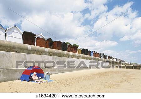 Stock Image - Man relaxing in small plastic tent on beach below row of beach hutsat Frinton in Essex. Fotosearch