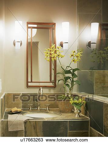 Stock Image - Mirror above yellow orchids on stone-tiled modern vanity unit. Fotosearch