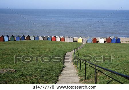 Stock Photo - Path across grassy field to row of brightly coloured beach huts by the blue sea. Fotosearch