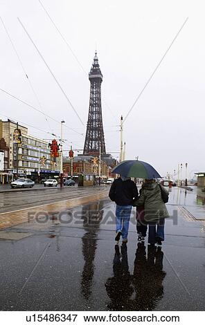 Stock Photo - People walking beneath umbrella in the rain at Blackpool with Blackpool Tower in the background. Fotosearch