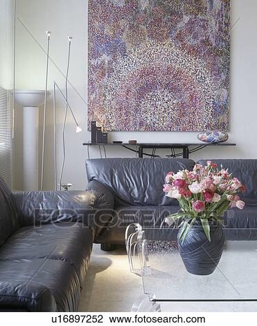 Stock Image - Pink tulips in vase on glass table in living room with large abstract painting and black leather sofas. Fotosearch