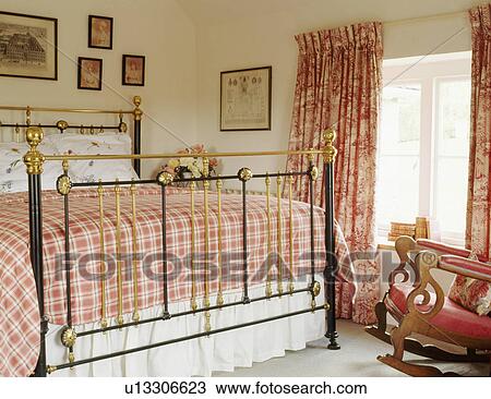 Stock Image - Red checked bedlinen on antique brass bed in country bedroom. Fotosearch