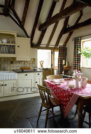 Stock Image - Red floral cloth on table in cream country kitchen with stone flooring and beamed apex ceiling . Fotosearch