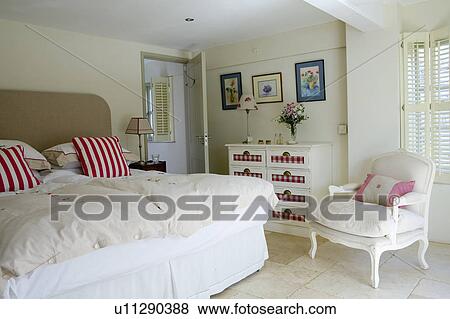 Stock Photo - REd striped cushions and cream duvet on bed in coastal bedroom ith white armchair in converted boathouse. Fotosearch