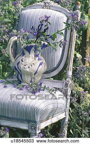 Stock Image - Still-life of blue agapanthus in antique blue+white textured jug on pale grey striped upholstered chair. Fotosearch