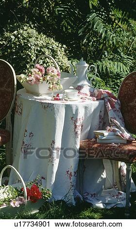 Stock Image - Still-life of pink roses in trug and white coffeepot on table with rose-patterned cloth in summer garden. Fotosearch