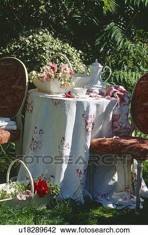 Stock Image - Still-life of pink roses in trug and white coffeepot on table with rose-patterned cloth in summer garden. Fotosearch