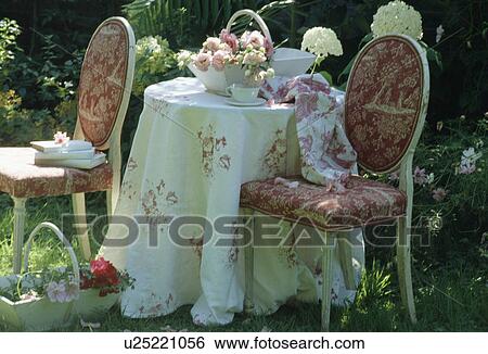 Stock Photograph - Still-life of rose-patterned cloth on table with patterned red chairs in summer garden. Fotosearch