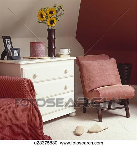 Stock Image - Sunflowers on white chest of drawers in attic bedroom with red upholstered chair against red wall. Fotosearch