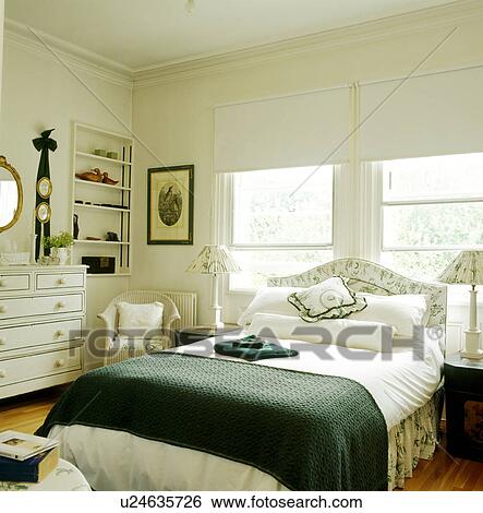 Stock Photograph - White blinds on windows behind bed with white linen and dark green throw. Fotosearch