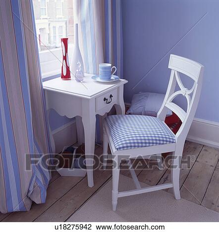 Stock Image - White chair and small table in front of window with blue+beige striped curtains. Fotosearch