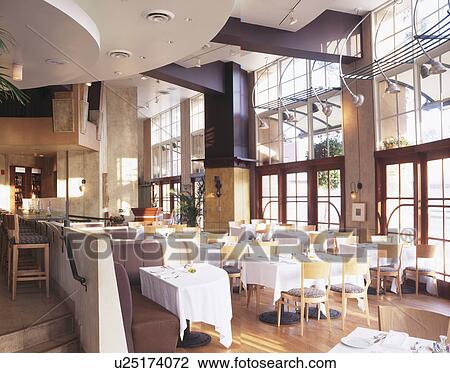 Stock Image - White cloths on table with pale wood chairs in modern double storey restaurant. Fotosearch