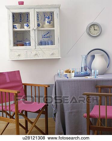 White Glass Front Cupboard On Wall Above Pink Director S Chairs And Table With Blue Cloth In Informal Dining Room Stock Photo U28121308 Fotosearch