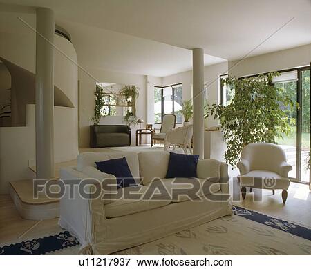 Stock Photo - White sofa and armchair in modern white openplan living room with tall houseplant beside patio doors. Fotosearch