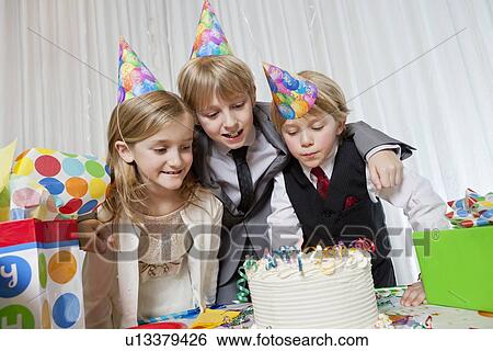 Stock Photograph - Brothers and sister wearing party hat looking at birthday cake together. Fotosearch