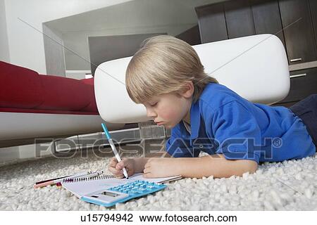 Stock Image - Elementary age boy writing in book while lying down on rug in house. Fotosearch