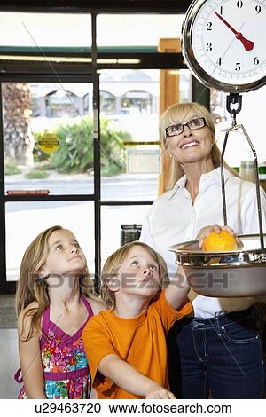 Stock Image - Happy woman and girl looking up while boy measuring fruit weight in market. Fotosearch