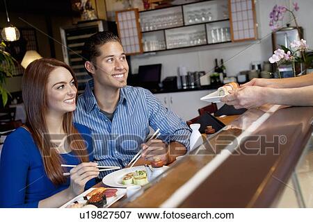 Stock Photo - Happy young couple looking at chef serving them sushi in restaurant. Fotosearch