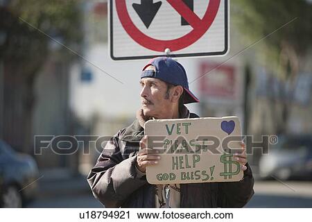 Stock Image - Homeless person with placard in front of road sign. Fotosearch